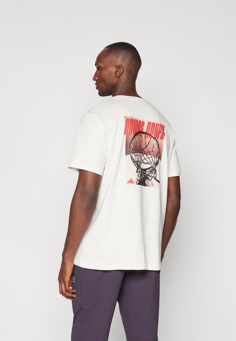 Tricou alb din bumbac cu un imprimeu grafic pe spate, care prezintă un coș de baschet și litere roșii. Mâneci scurte și croi lejer.