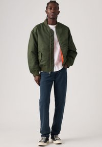 Casaco bomber verde com punhos e gola canelados, apresenta fecho de correr frontal e dois bolsos laterais. Usado sobre uma t-shirt branca e jeans azuis.