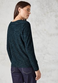 Dunkelblauer Pullover mit strukturiertem Strick, runder Ausschnitt und langen Ärmeln. Kombiniert mit dunklen Jeans, die eine lockere Passform und subtile Details zeigen.