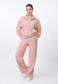 Set de îmbrăcăminte confortabilă roz din fleece, format dintr-un hanorac cu fermoar și guler înalt și pantaloni evazați. Textură moale și croială lejeră.