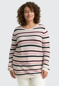 Lachende vrouw met krullend haar die een lange mouw geribbelde trui draagt met horizontale rozenkleurige, zwarte, witte en bordeauxrode strepen.