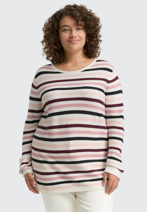 Lachende vrouw met krullend haar die een lange mouw geribbelde trui draagt met horizontale rozenkleurige, zwarte, witte en bordeauxrode strepen.