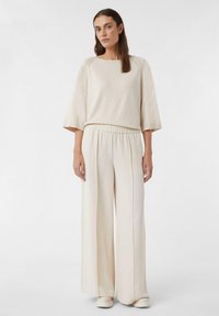 Beige gestricktes Langarmoberteil mit rundem Ausschnitt, kombiniert mit beigen weit geschnittenen Hosen mit elastischem Bund, getragen mit passenden Schuhen.
