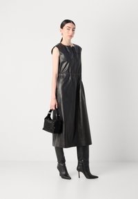 Schwarzes Lederkleid mit taillierter Taille, ärmellosem Design und Rundhalsausschnitt. Kombiniert mit knielangen, spitzen schwarzen Stiefeln und einer kleinen schwarzen Handtasche.