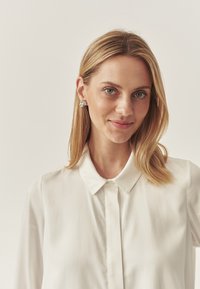 Biała bluzka z kołnierzykiem, gładką fakturą i zapinana z przodu na guziki. Modelka ma na sobie srebrne geometryczne kolczyki. Tło z miękkim oświetleniem.