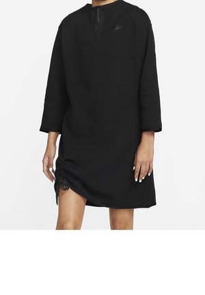 Robe noire à manches longues avec une courte fermeture éclair à l'avant, logo Nike sur la poitrine, et ourlet froncé ajustable sur la cuisse gauche portée par un mannequin.