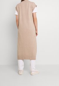 Béžové pletené šaty bez rukávů s žebrovaným lemem, nošené přes bílé tričko a doplněné krémovými teniskami, s dlouhým, uvolněným střihem.