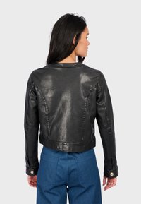 Černá kožená bunda s krčeným střihem, zdobená šitými detaily a knoflíkovými manžetami, nošená přes modré kalhoty s vysokým pasem.