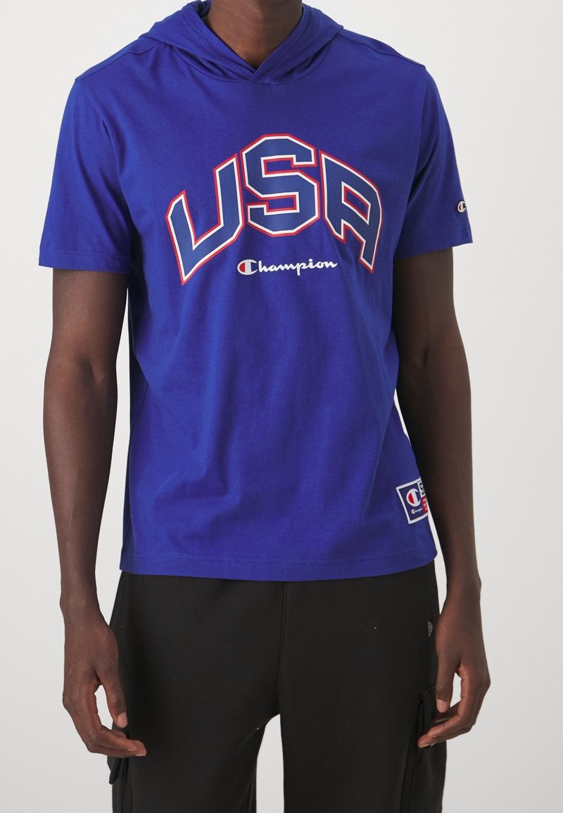 Niebieska bluza z krótkim rękawem z napisem "USA" w czerwonych i białych literach, z logo Champion. Wykonana z gładkiego materiału w luźnym kroju.