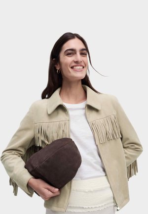 Giacca in suede beige con frange sul petto, abbinata a una maglietta bianca e una borsa marrone texturizzata tenuta nella mano sinistra.