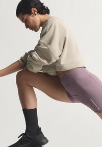 Αθλητική εμφάνιση με ανοιχτό μπεζ, φαρδύ φούτερ και εφαρμοστά λιλά σορτς. Μαύρες κάλτσες μέχρι το γόνατο και αθλητικά μαύρα παπούτσια.