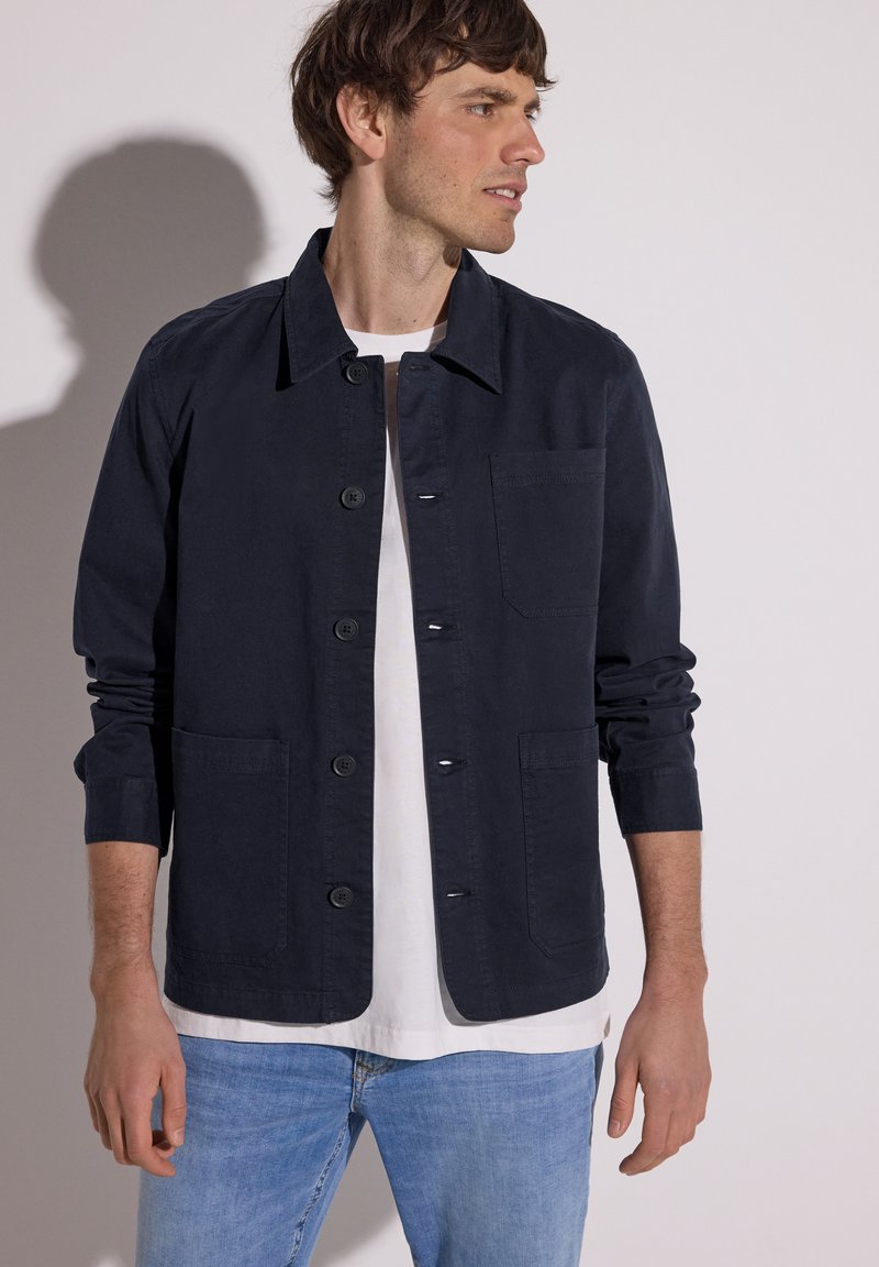 Mann trägt eine marineblaue Jacke mit Knöpfen über weißem T-Shirt und hellblauen Jeans, blickt nach rechts vor einfachem Hintergrund.