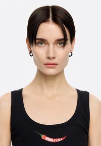 Débardeur noir à côtes avec un graphique de piment rouge et "BIMBA Y LOLA." Le modèle porte des boucles d'oreilles en perles. Peau lisse, cheveux courts et foncés.