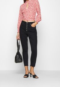 Black high-waisted jeans with a five-button front, fitted design, and frayed hems, paired with a floral red blouse and black accessories.