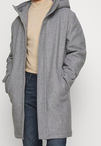 Grå ullblandad kappa med huva, som har tryckknappar, sidofickor och en avslappnad passform. Bärs över en beige polotröja och mörka jeans.