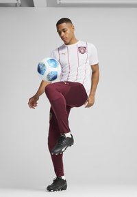Camiseta branca da Puma com detalhes em bordô e logotipo, combinada com calças fitted bordô e sapatilhas pretas, equilibrando uma bola de futebol azul com padrão.