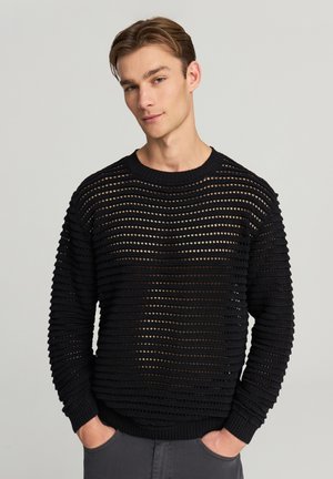 Giovane uomo con le mani in tasca che indossa un maglione nero a maglia aperta su una maglietta più scura, in piedi contro uno sfondo chiaro e semplice.