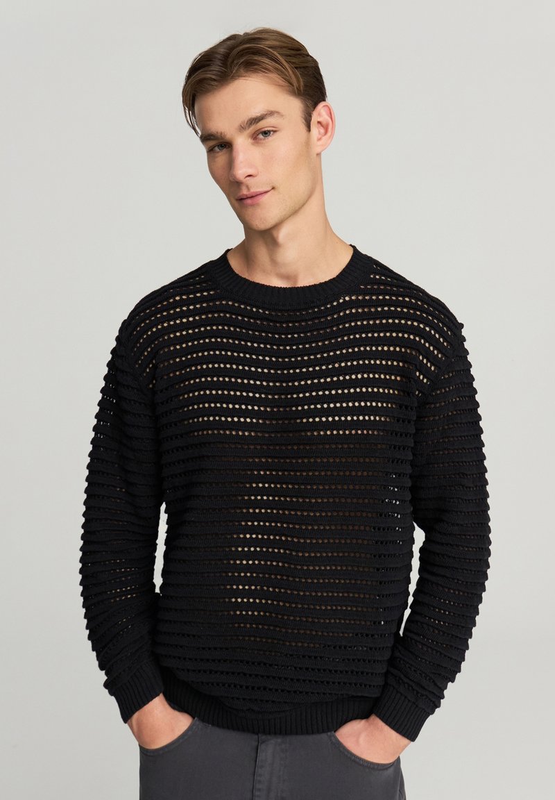 Giovane uomo con le mani in tasca che indossa un maglione nero a maglia aperta su una maglietta più scura, in piedi contro uno sfondo chiaro e semplice.