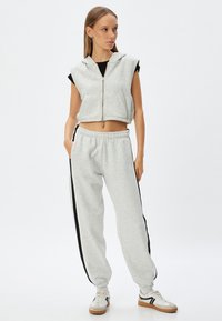 Lichtgrijze cropped hoodie met rits en capuchon, gecombineerd met lichtgrijze sweatpants met zwarte zijstrepen. Model draagt sneakers.