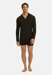 Tmavě zelená košile s dlouhým rukávem, límečkem a knoflíky, v kombinaci s černými šortkami. Materiál vypadá měkký a přiléhavý. Bosý.