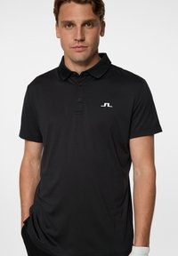 Mustapaita, jossa on kaulus, lyhyet hihat ja kolme nappia. Rinnassa valkoinen logo ja sileä, teksturoitu kangas.