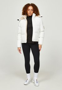 Witte pufferjack met een zachte beige voering, ritssluiting en staande kraag, gedragen over een zwart longsleeve top en zwarte leggings.