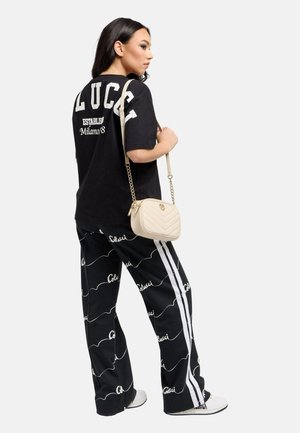 Kvinde set fra siden iført sort oversized T-shirt med hvid tekst, sorte mønstrede bukser med hvide striber, hvide sneakers og beige skuldertaske.