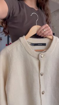 Personne tenant un cardigan beige boutonné sur un cintre en bois dans un cadre intérieur, avec des cheveux bouclés et une chemise sombre visibles.
