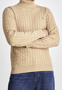 Beige Rollkragenpullover aus Kabelstrick mit gerippten Bündchen und Saum. Verfügt über ein strukturiertes Muster und ein kleines Logodetail auf der Brust.