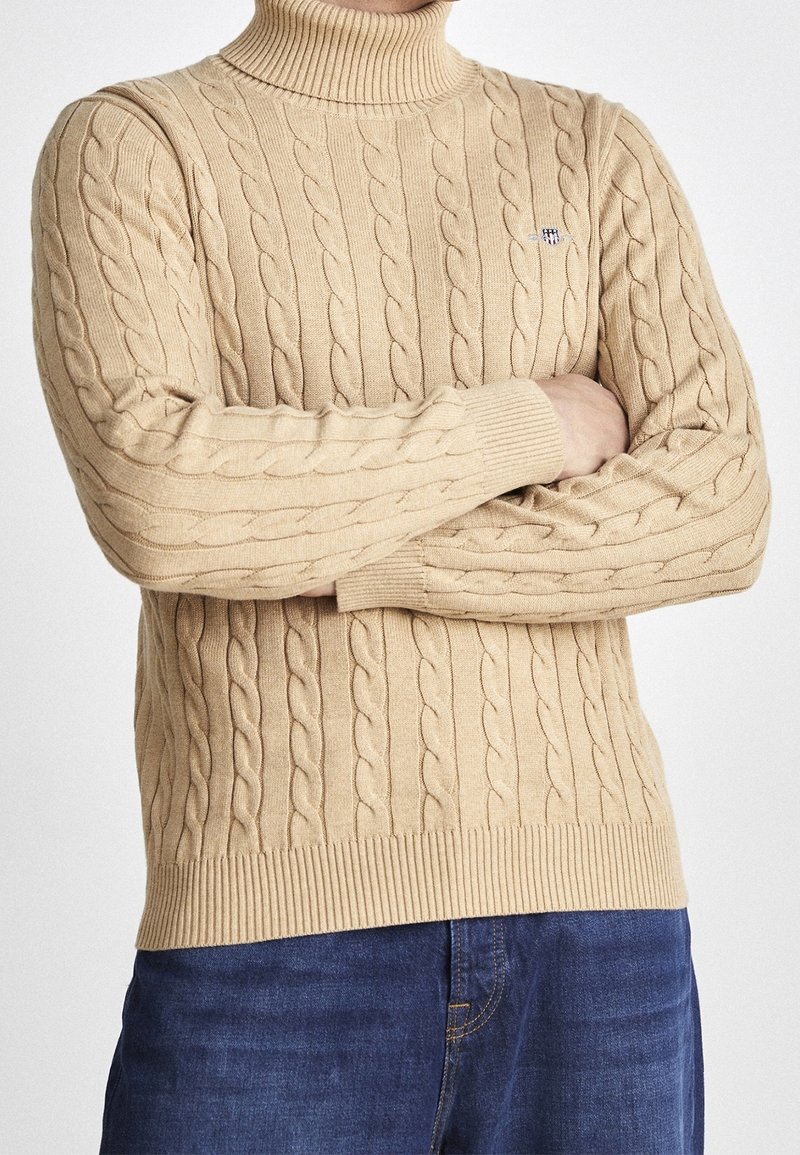 Beige Rollkragenpullover aus Kabelstrick mit gerippten Bündchen und Saum. Verfügt über ein strukturiertes Muster und ein kleines Logodetail auf der Brust.