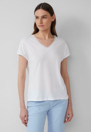 T-shirt blanc à manches courtes avec un décolleté en V orné d'une délicate bordure en dentelle. Texture douce et lisse, coupe décontractée. Associé à un pantalon bleu clair.