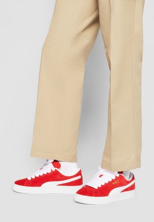 Czerwone zamszowe sneakersy z białymi akcentami, z grubą białą podeszwą i białymi sznurówkami, w zestawie z beżowymi spodniami.