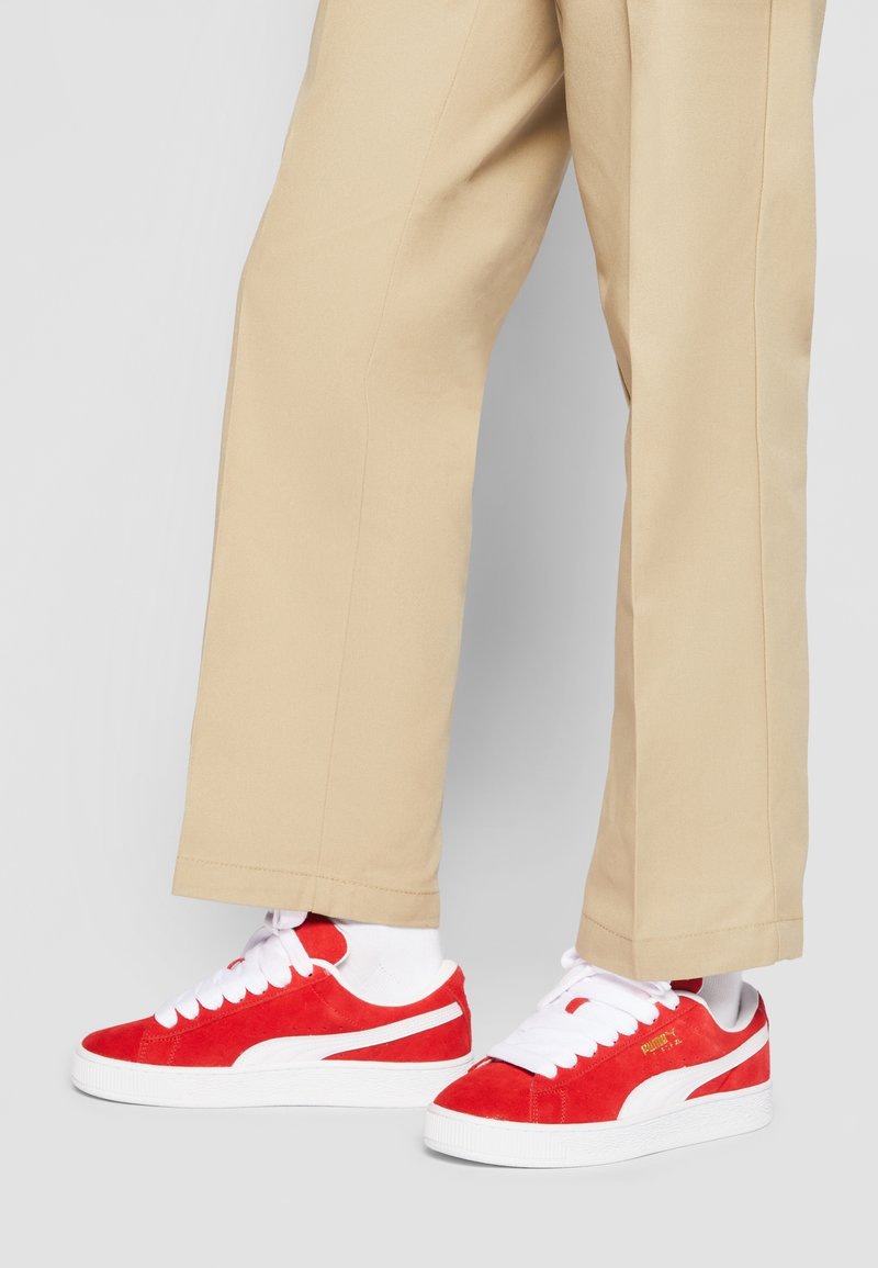 Red suede sneakers with white accents, featuring a thick white sole and white laces, paired with beige trousers.