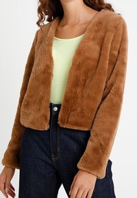 Giacca corta in pelliccia sintetica marrone con scollatura a V, maniche lunghe e texture morbida, abbinata a un top verde chiaro e jeans in denim scuri.