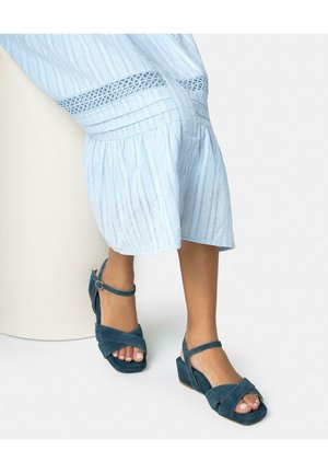 Persoon draagt lichtblauwe gestreepte wijde broekspijpen en teal suède open teen enkellaarzen met sleehak, staand tegen een witte achtergrond.