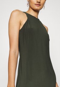 Žena v bezramínkových tmavě zelených přiléhavých šatech s vysokým výstřihem a malými záhyby na ramenních švech, doplněná visícími náušnicemi.