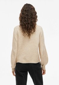 Femme aux longs cheveux bouclés bruns, portant un pull beige et un pantalon noir, se tenant de dos devant un fond blanc.