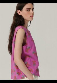 Jeune femme aux longs cheveux foncés portant un haut sans manches rose à motifs de cerises rouges, vue de profil sur un fond uni.