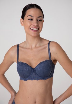 Giovane donna sorridente, indossa un reggiseno di pizzo blu scuro, con le mani appoggiate sui fianchi davanti a uno sfondo semplice.