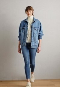 Giacca di jeans con due tasche frontali, indossata sopra un maglione a collo alto bianco a coste. Abbinata a jeans scuri e sneakers beige.