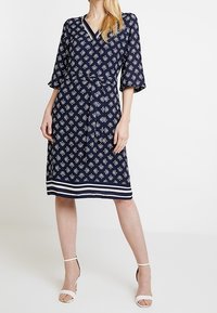 Mörkblå klänning med vitt blommönster, V-ringning, trekvartslånga ärmar, åtsittande midja och randig fåll; i kombination med vita klacksandaler.