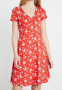 Punainen mekko V-pääntiellä, lyhyillä hihoilla ja istuvalla bodysuitilla. Siinä on valkoinen kukkakuosi ja levenevä hameosa. Sileä kangas.