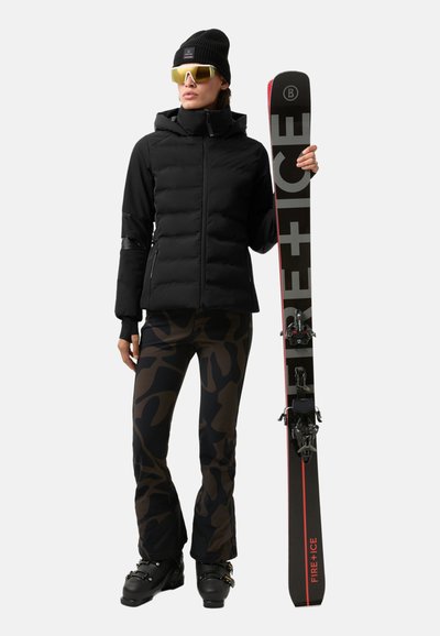 Zwarte donsjas, camouflagebroeken met patroon, skischoenen en een ski met een zwart-rood ontwerp. Accessoires zijn onder andere een zwarte muts en gele zonnebril.