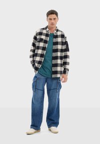 Camisa de franela a cuadros en blanco y negro sobre una camiseta teal, combinada con pantalones cargo azules. Incluye bolsillos y tiene un corte relajado.