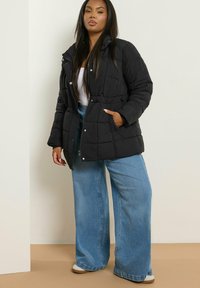 Doudoune noire matelassée avec zip sur le devant, poches latérales et taille ajustable avec cordon, associée à un jean en denim à jambes larges et des baskets.