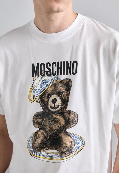 Balta medvilninė marškinė su raštu, vaizduojančiu lokį mėlyname ir baltame inde, uždengtame dekoratyvine taure, su „Moschino“ prekės ženklu.
