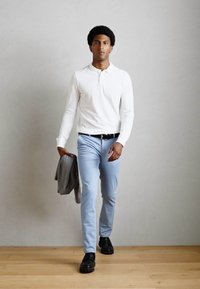 Weißes Langarmpolo, hellblaue Hose, schwarze Schuhe; hält eine graue Jacke; steht auf einem Holzboden vor einer einfarbigen Wand.