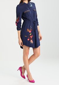 Mujer con un vestido azul marino con mariposas y flores bordadas, cintura ceñida y zapatos de tacón alto rosas brillantes, de pie contra un fondo blanco.