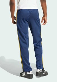 Marinblå träningsbyxor med gula sidostreck, tillverkade av ett slätt tyg. Har resårmidja och en figurnära passform; använda med svarta sneakers.