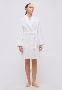 Γυναίκα που στέκεται ξυπόλητη φορώντας ένα λευκό μπουρνούζι μέσης μήκους με γιακά σαν σάλι και ζώνη, μπροστά σε απλό φόντο.